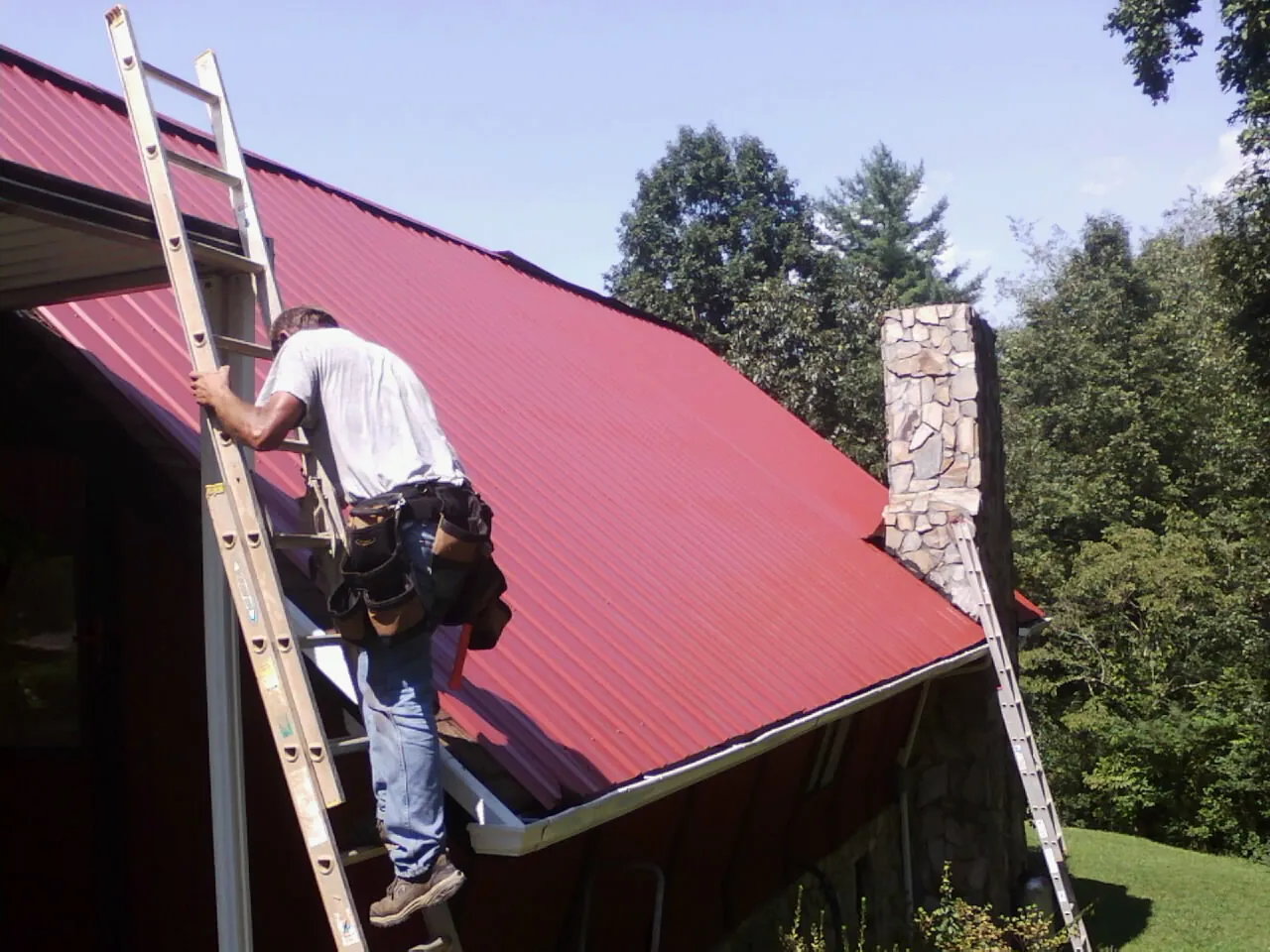 Roofer on a ladder beside a red metal roof and stone chimney — Soffit & Fascia Repair by Roofing Brothers in Ben Lomond